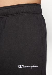 Pantalons de survêtement noirs en tissu mélangé de coton, avec une taille élastique et un logo "Champion" brodé en blanc sur le côté.