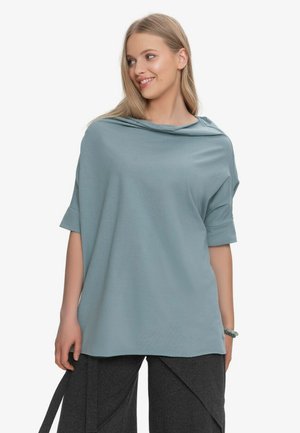 Młoda kobieta patrzy bokiem, ubrana w luźną jasnoniebieską bluzkę z podwiniętymi rękawami i ciemne szerokie spodnie z nakładającymi się panelami.