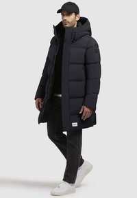 Mørkeblå pufferjakke, knælang med hætte, med lynlås, elastisk snor og mærkedetalje. Sammen med sorte jeans og hvide sneakers.