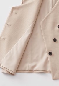 Blazer beige ouvert montrant une doublure intérieure lisse avec des boutonnières sur le côté gauche et trois boutons foncés sur le côté droit.
