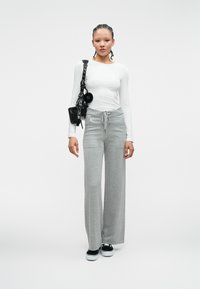 Biała koszula z długim rękawem, szare szerokie spodnie dresowe z troczkiem, czarna torba na ramię i czarne sneakersy na jednolitym tle.