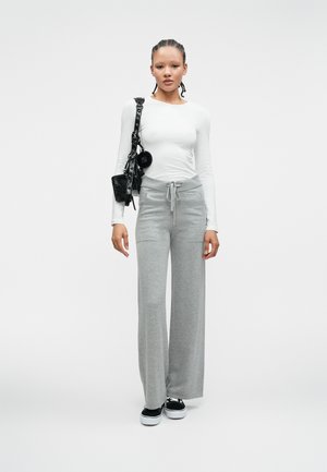 Biała koszula z długim rękawem, szare szerokie spodnie dresowe z troczkiem, czarna torba na ramię i czarne sneakersy na jednolitym tle.