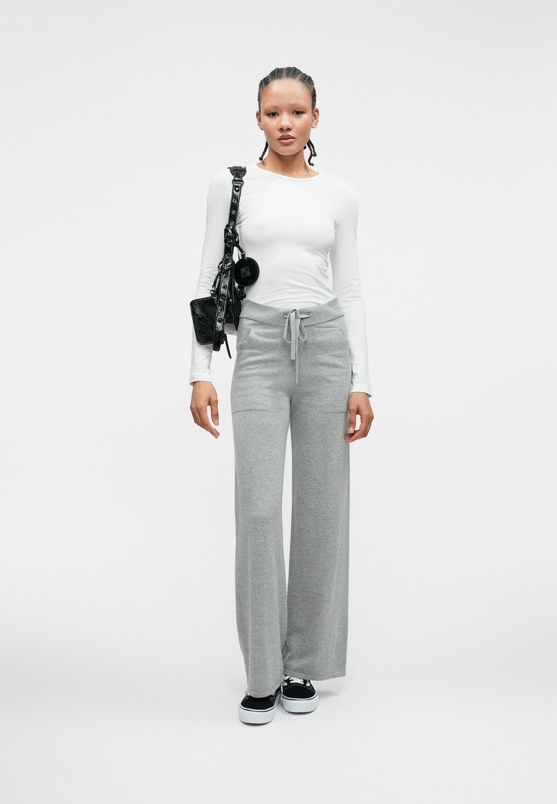 Biała koszula z długim rękawem, szare szerokie spodnie dresowe z troczkiem, czarna torba na ramię i czarne sneakersy na jednolitym tle.