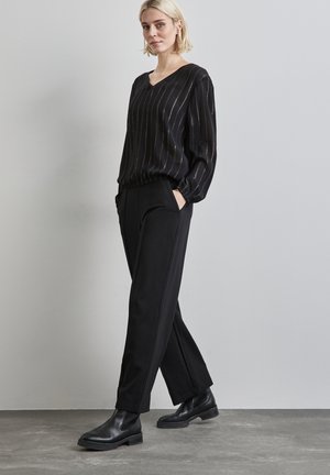 Schwarze Bluse mit goldenen vertikalen Streifen, V-Ausschnitt und elastischem Bund. Schwarze Tailleurhose mit Taschen, kombiniert mit schwarzen Stiefeletten.
