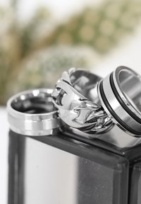 Four silver rings stacked, featuring smooth, woven, and grooved designs. The background is blurred, highlighting the rings' details.