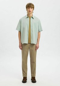 Korte mouwen, groen en wit gestreept shirt met een knoopsluiting, gecombineerd met een bruine T-shirt en beige broek. Bruine schoenen maken de outfit compleet.