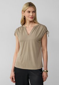 Haut beige à manches courtes avec un col en V, des détails froncés aux épaules et des liens. Tissu doux avec une texture lisse. Associé à un pantalon noir.