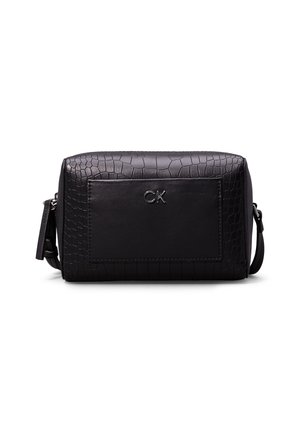 Trousse de toilette en cuir noir texturé avec poche avant et logo argenté "CK", forme rectangulaire et bandoulière réglable sur fond blanc.