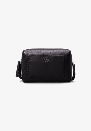Trousse de toilette en cuir noir texturé avec poche avant et logo argenté "CK", forme rectangulaire et bandoulière réglable sur fond blanc.