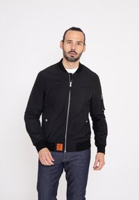 Svart bomberjacka med framdragkedja, ribbad ärm, och en orange etikett; har sidofickor och ett slitstarkt, lätt tyg.