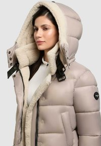 Beige Pufferjacke mit einem flauschigen cremefarbenen Futter, großer Kapuze, Druckknöpfen und Seitentaschen. Verfügt über ein Logo am Ärmel.