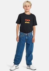 Schwarzes T-Shirt mit rot-gelbem Wellenmuster, kombiniert mit weit geschnittenen blauen Jeans und weißen Turnschuhen mit grünen Akzenten.