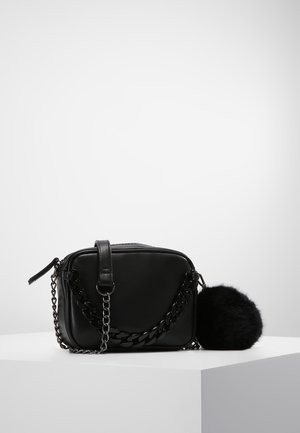 Schwarze rechteckige Umhängetasche mit klobigem Kettenriemen und angebrachtem schwarzem flauschigem Bommel, liegend auf einem weißen Podest.