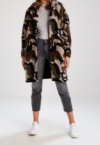 Camo-mønstret fleecefrakke i jordfarver, knaplukning foran, oversized design, parret med grå jeans og hvide sneakers.