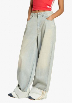Persona che indossa jeans a gamba larga color azzurro chiaro con risvolti arrotolati e una maglietta corta rossa Adidas, con le mani nelle tasche, in piedi davanti a uno sfondo bianco.