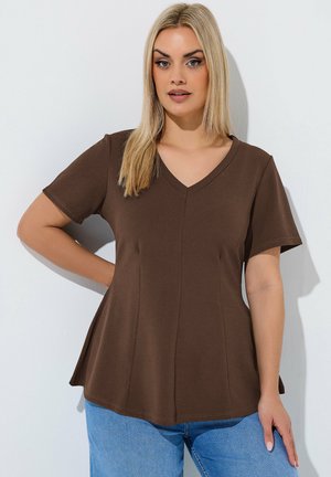 Kvinde med glat blond hår iført en brun V-hals kortærmet top og blå jeans, der står mod en hvid baggrund.