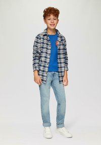 Junge trägt ein blaues T-Shirt mit Grafik, ein offenes kariertes Hemd in Marineblau und Weiß, hellblaue Jeans und weiße Turnschuhe. Lockige rötliche Haare.