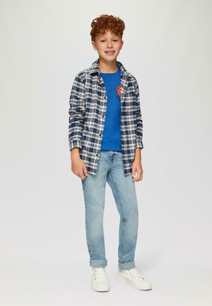 Junge trägt ein blaues T-Shirt mit Grafik, ein offenes kariertes Hemd in Marineblau und Weiß, hellblaue Jeans und weiße Turnschuhe. Lockige rötliche Haare.