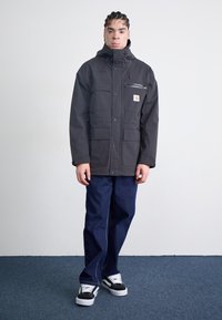 Zwarte, waterdichte jas met een capuchon, meerdere voorzakken en een ritszak op de borst. Draagt met donkerblauwe broek en zwarte sneakers.