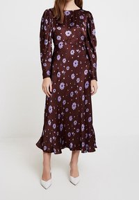 Brun blommig midiklänning med långa puffärmar, prydd med lila blommor och en svängande nederkant. Kombinerad med vita spetsiga skor.
