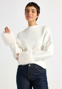 Kvinde iført hvid sweater med fluffy manchetter og mørkeblå jeans, poserende med den ene hånd hævet nær skulderen mod en ensfarvet baggrund.