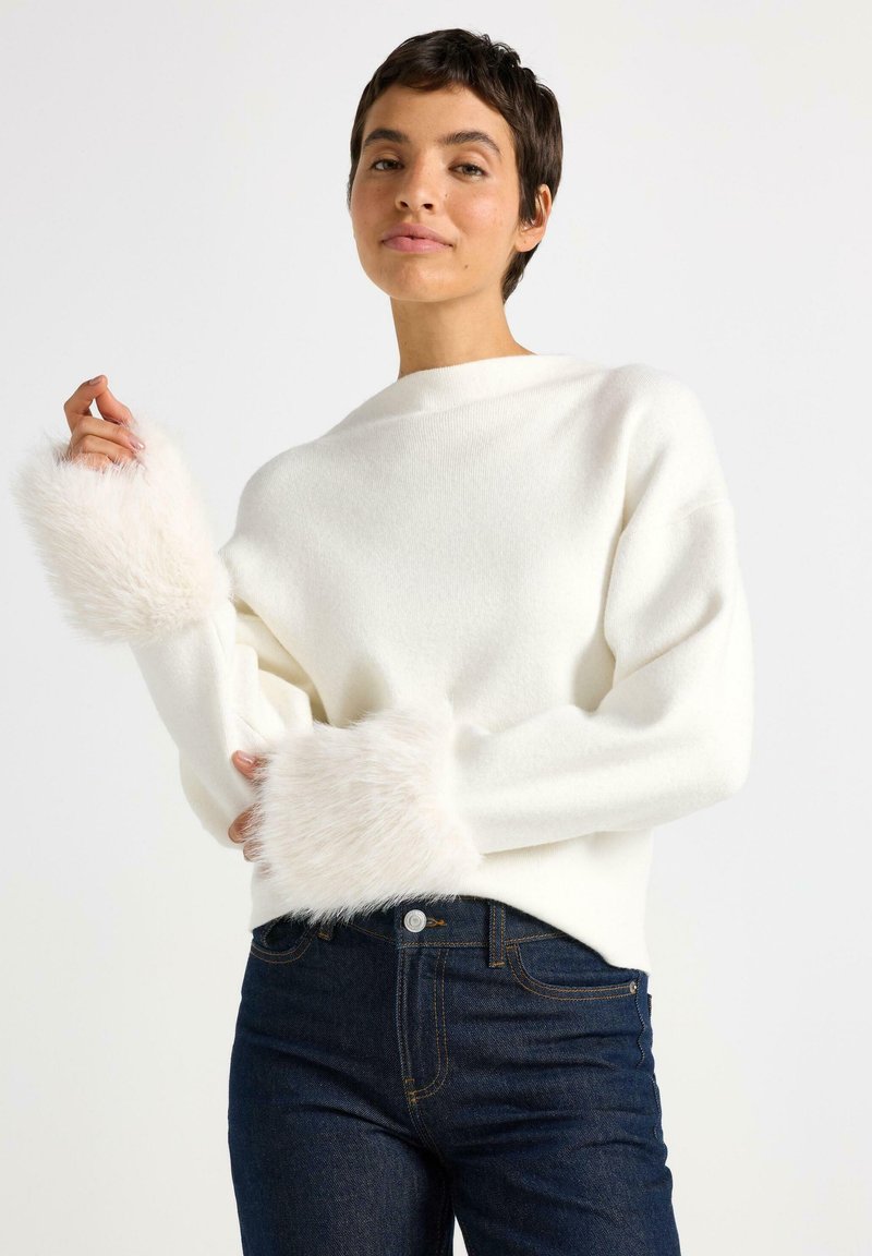 Kvinde iført hvid sweater med fluffy manchetter og mørkeblå jeans, poserende med den ene hånd hævet nær skulderen mod en ensfarvet baggrund.