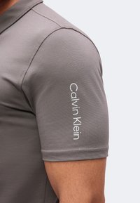 Gros plan sur le haut du bras d'une personne portant un t-shirt gris à manches courtes avec le nom de la marque Calvin Klein imprimé verticalement sur la manche.
