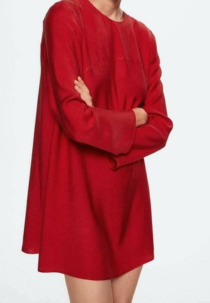Personne portant une robe rouge ample à manches longues larges, debout les bras croisés devant un fond clair uni.