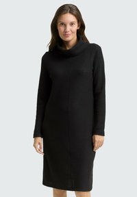 Robe en maille noire avec col bénitier, manches longues et silhouette droite. Tissu texturé avec des détails subtils et des fentes latérales à l'ourlet.