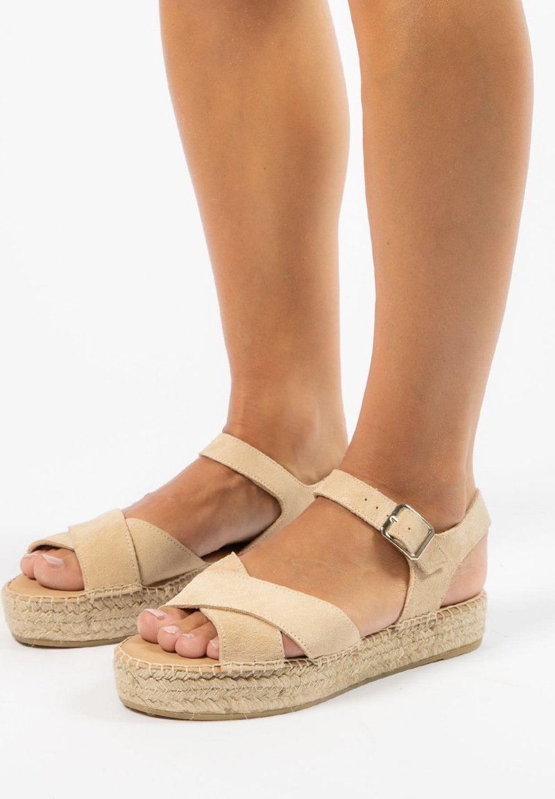 Beige suède sandalen met gekruiste banden, enkelgesp en een geweven jute platform. Open teen ontwerp dat de tenen met een natuurlijke afwerking tentoonstelt.