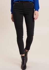 Jean skinny noir avec poches devant, détails en métal argenté et léger stretch. Porté avec des bottes noires à chevilles, montrant des zips aux chevilles.