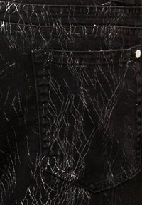 Jeans in denim nero con un motivo bianco screpolato, dotati di una tasca posteriore e dettagli in hardware argentato. La texture appare liscia e leggermente lucida.