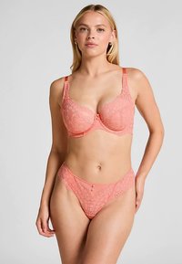 Kanten lingerieset in koraal, met een balconette beha met decoratieve strik en bijpassende string. Geheel voorzien van textuur in kant.