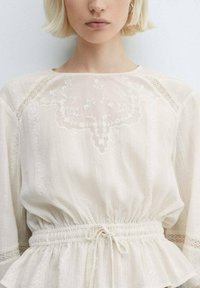 Blusa bianca in tessuto testurizzato, con dettagli in pizzo e vita arricciata. Maniche lunghe e morbide con accenti in pizzo.