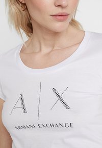 Balta trumpa rankovėmis marškinėliai su juodais rhinestone akcentais, sudarančiais "A/X" ir "Armani Exchange" tekstus, švelni medvilnė ir apvalus iškirpimas.