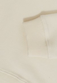 Kreemi värvi kudumisega sviiteri varrukas koos soonikuga detailiga tekstuuriga kangas taustal.