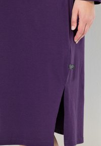 Lila bomullsklänning med en sidospalt och en vävd etikett. Mjuk textur, korta ärmar och rak fåll med detaljer i botten.