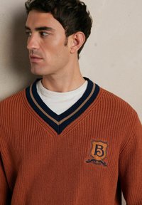 Sweter w serek z pomarańczowego dzianinowego materiału z granatowym i beżowym paskiem przy kołnierzu. Posiada haftowane logo na piersi. Gładka faktura.