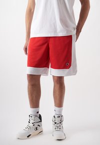 Rote Basketballshorts mit weißen Mesh-Akzenten, strukturiertem Stoff und einem Logopatch. Kombiniert mit weißen Turnschuhen und gemusterten Socken.