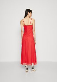 Rochie maxi roșie cu bretele subțiri și un strat fluid de voal. Are un corset ajustat și o textură netedă. Este purtată cu botine cu toc argintii strălucitoare.
