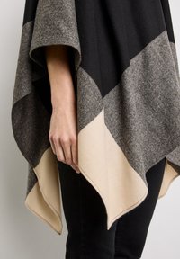 Poncho i sort, grå og beige uldblanding; har geometrisk farveblokering, blød tekstur, og et flydende design med en asymmetrisk kant.