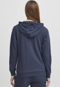 Tumši zilas krāsas kapuciņa džemperis, kas izgatavots no mīksta auduma, ar brīvu piegriezumu, garām piedurknēm un ribotām aprocēm, kā arī ar kangaru kabatu priekšpusē.