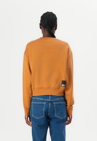 Afgekorte, mosterdgele sweatshirt van zacht materiaal met geribde manchetten en zoom. Bevat een zwart label op de achterkant. Gepaard met blauwe spijkerbroeken.