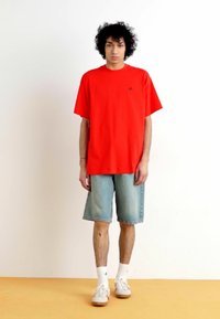 Tricou oversized roșu cu mâneci scurte, asortat cu pantaloni scurți din denim de culoare albastru deschis. Modelul poartă șosete albe și adidași bej, stând pe un podea portocalie.