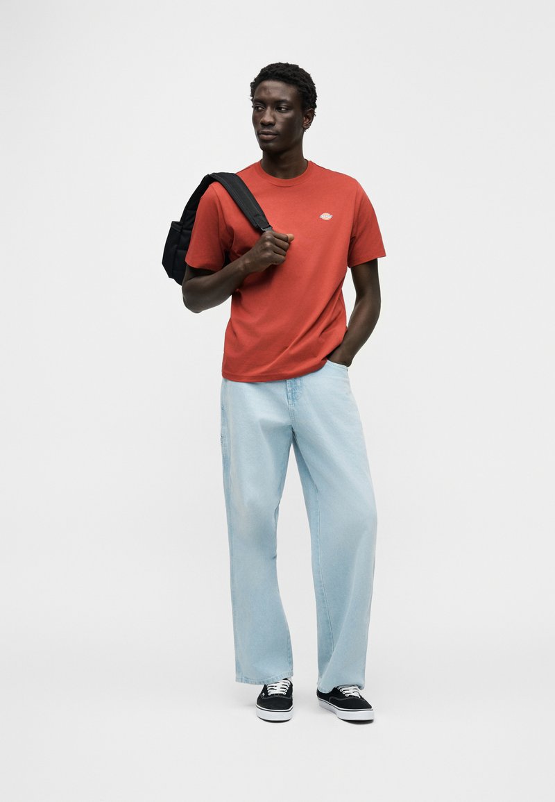 Rood katoenen T-shirt met een klein logo, gecombineerd met lichtblauwe wijde denim jeans. Zwarte rugzak en zwarte sneakers maken de look compleet.