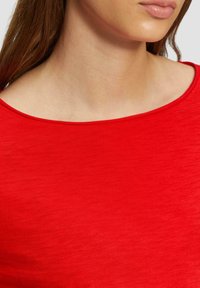 Haut rouge à encolure dégagée en tissu doux, présentant une texture lisse et un minimum de détails de couture le long de l'encolure.