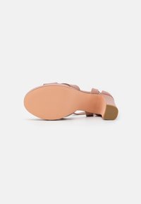 Sandale beige à talon bas couchée sur le côté, montrant une semelle lisse et un talon bloc épais sur fond blanc.