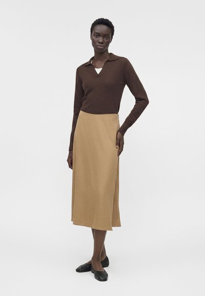 Γυναίκα όρθια, φοράει καφέ μπλούζα με μακριά μανίκια, μπεζ μίντι φούστα και μαύρα επίπεδα παπούτσια μπροστά σε απλό ανοιχτό φόντο.