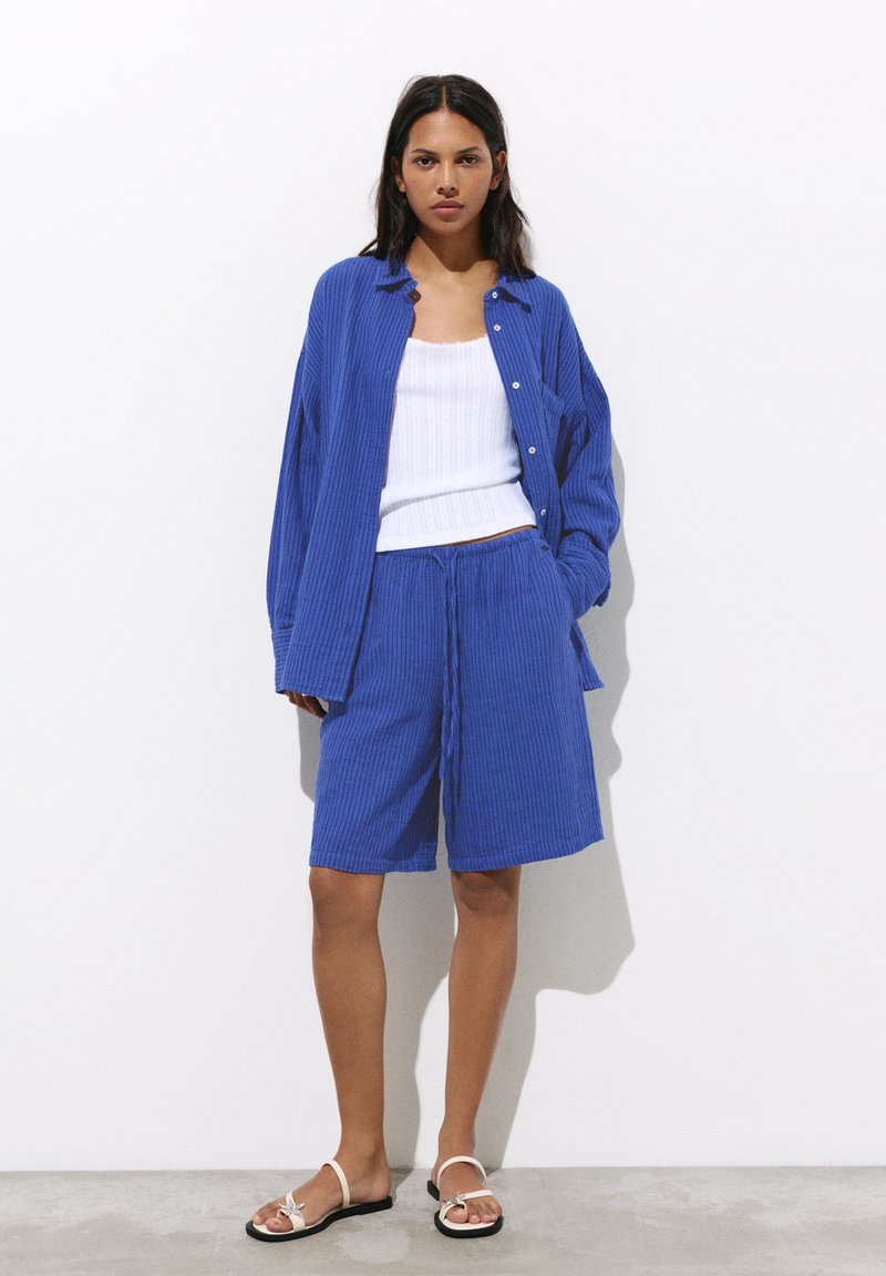 Femme portant une chemise bleue côtelée boutonnée et un short assorti avec un débardeur blanc et des sandales blanches plates, debout devant un fond blanc.