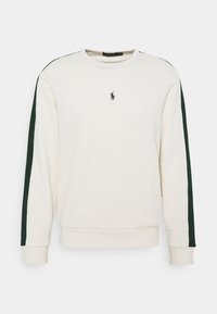 Krémová mikina z měkké látky, s tmavě zelenými pruhy na bocích a malým vyšívaným logem na hrudi. Kulatý výstřih a žebrované manžety.